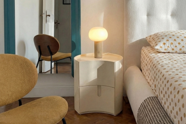 שידת לילה נאפו (Napo) מעוגלת בצבע שמנת, עליה מנורת שולחן מוארת, בחדר שינה עם כיסא חרדל ומיטה עם מצעים מנוקדים.