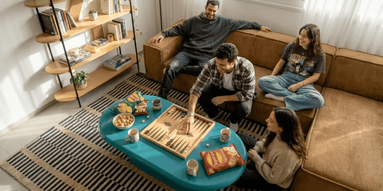 חברים משחקים בשמחה שש-בש בערב משחק בבית, יושבים על ספת Napo (נאפו) מודולרית בצבע קאמל (קורדרוי) וסביב שולחן סלון בגוון טורקיז מבריק. על השולחן כיבוד וספר.